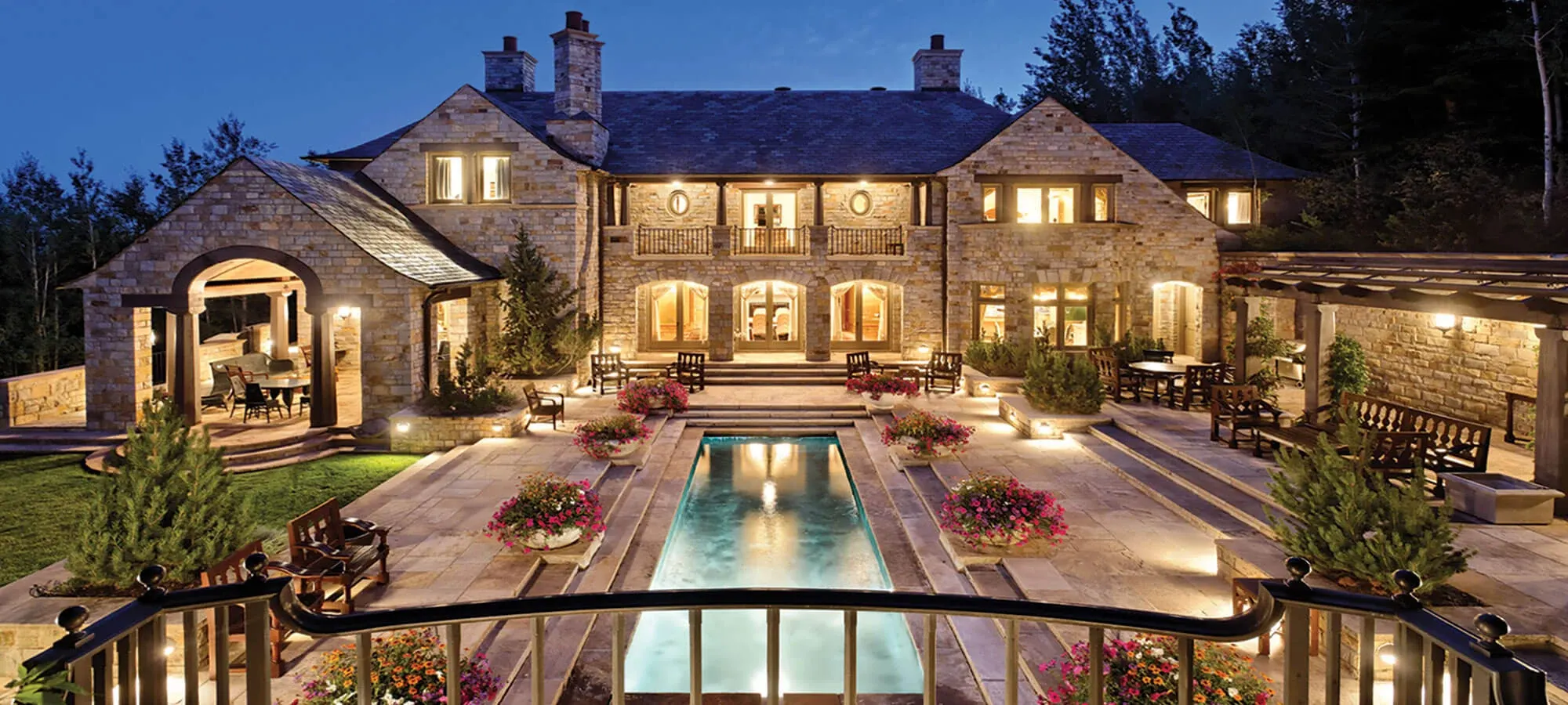 Luxury stone mansion at dusk with illuminated lap pool, manicured grounds, and multiple patios